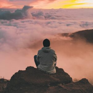 Person sitting on mountain peak, gazing at clouds