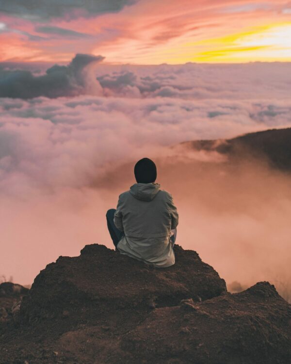 Person sitting on mountain peak, gazing at clouds